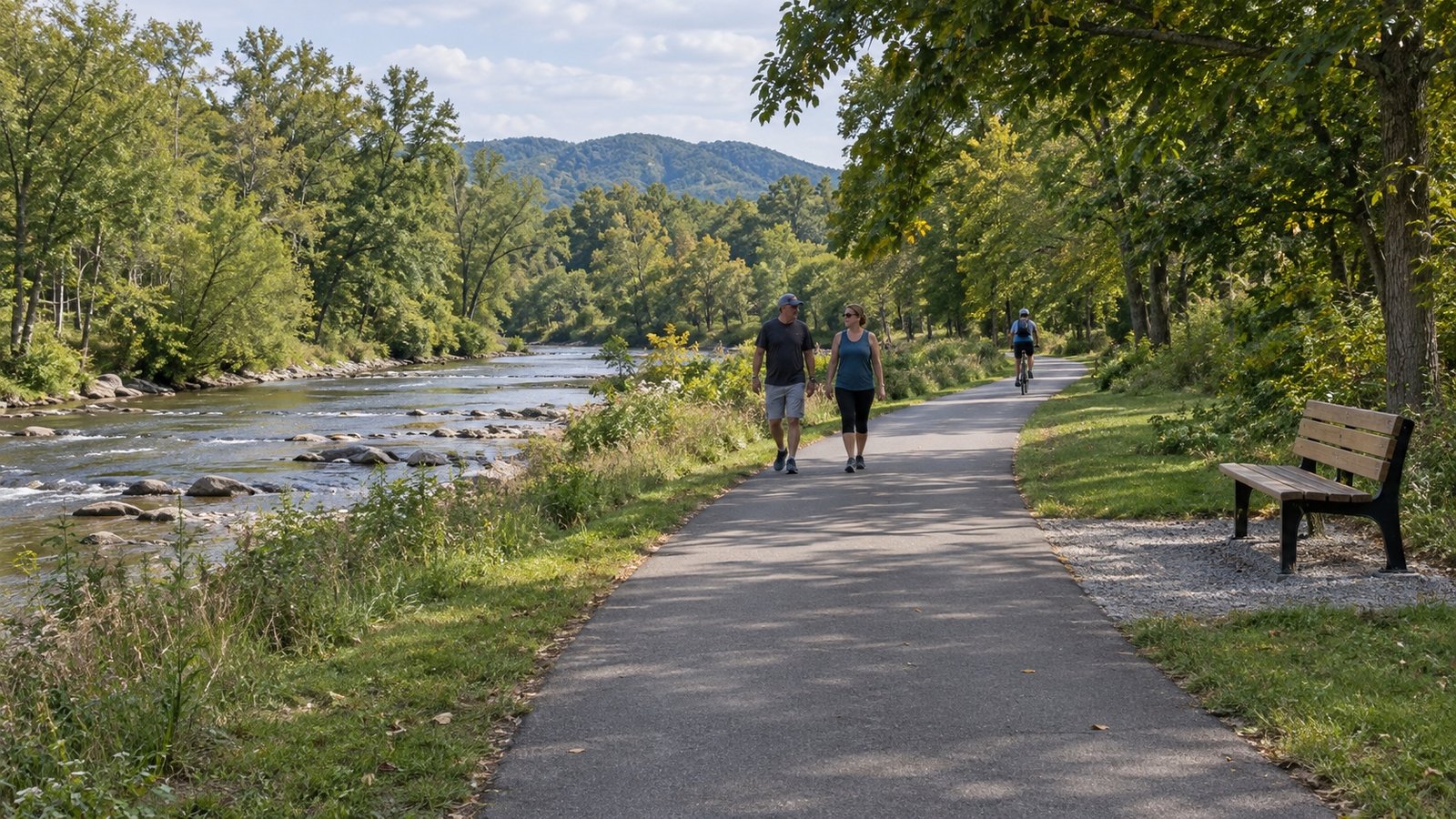 Two adults walking on a paved riverside path, with a cyclist farther away and a bench beside the path.