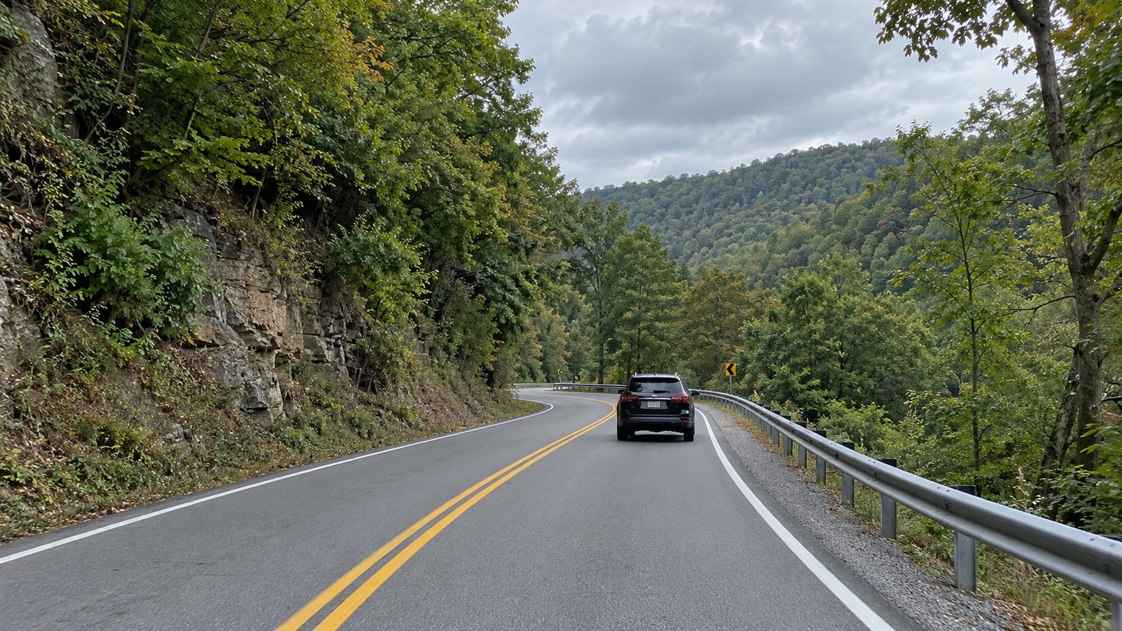 A whole SUV travels away in the right lane on a curved road through wooded hills.