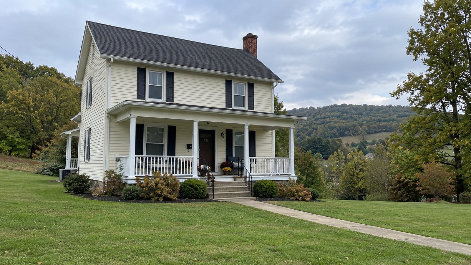 A whole cream two-story house with a front porch, lawn and wooded hills behind.