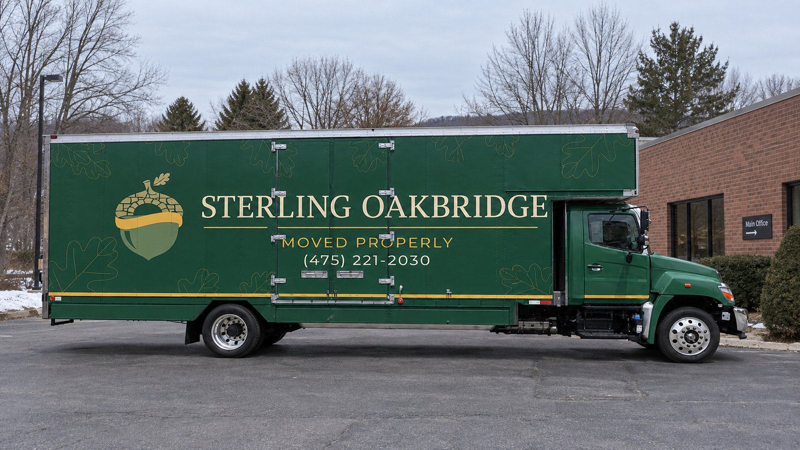 A complete green Sterling Oakbridge Hino box truck viewed side-on on asphalt beside a low brick office building