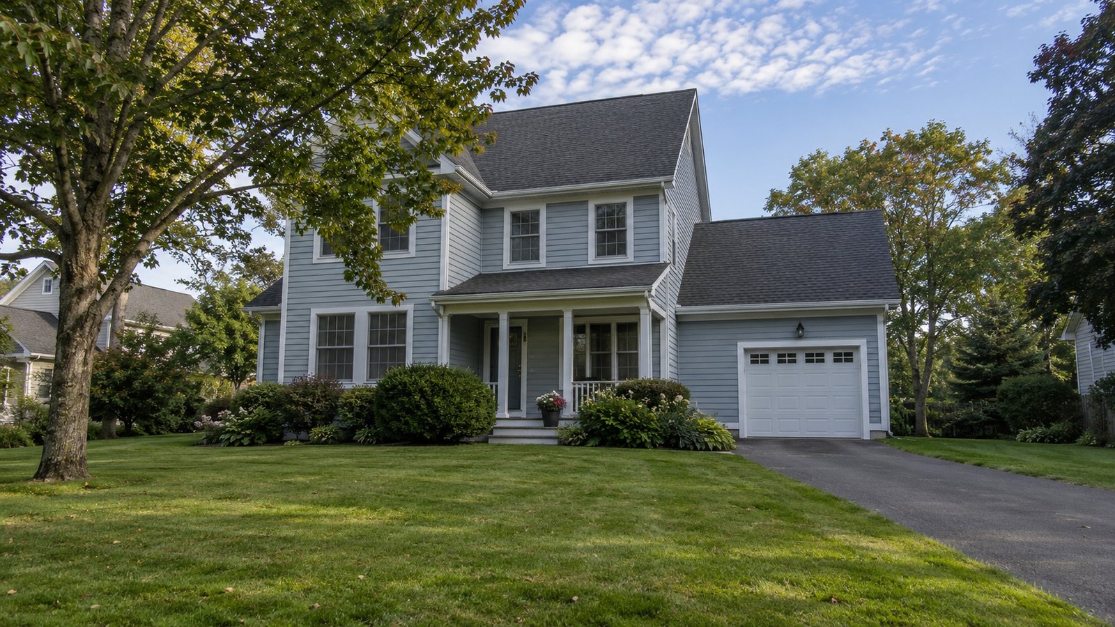 A blue-gray two-story clapboard house with a covered porch and attached garage on a broad lawn among mature trees