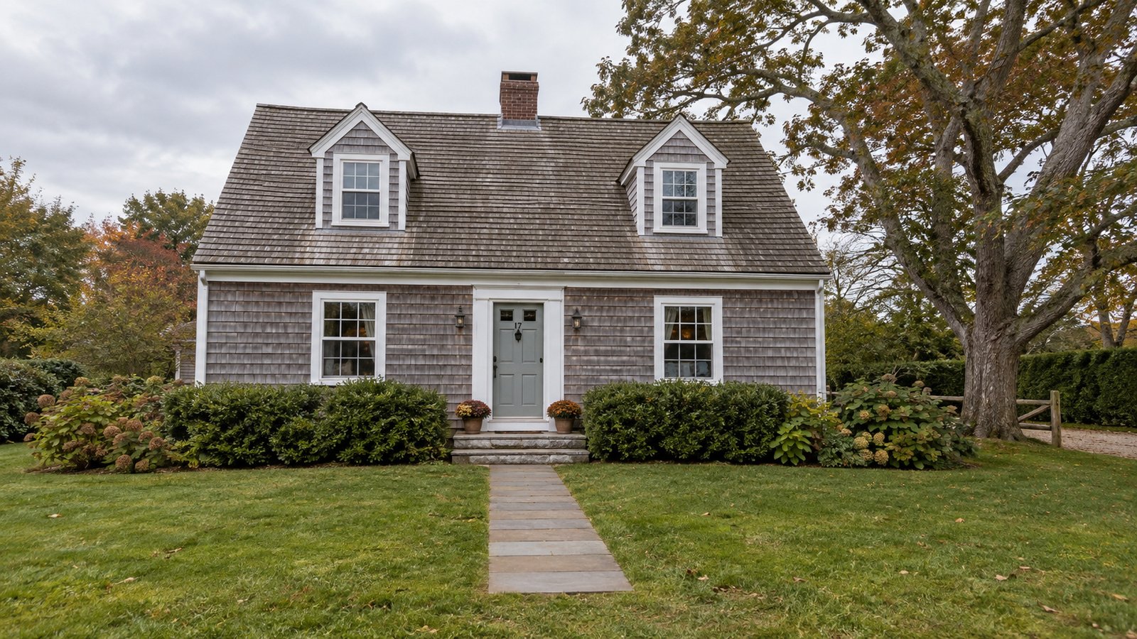 A modest cedar-shingle cottage Cedar-shingle cottage with two dormers, a central front door and a stone path across the lawn.