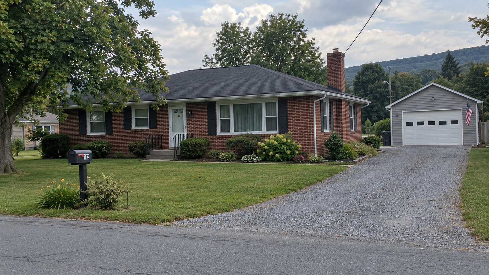 The house and its approach A modest red-brick ranch, separate gray garage and gravel driveway meeting a village street.