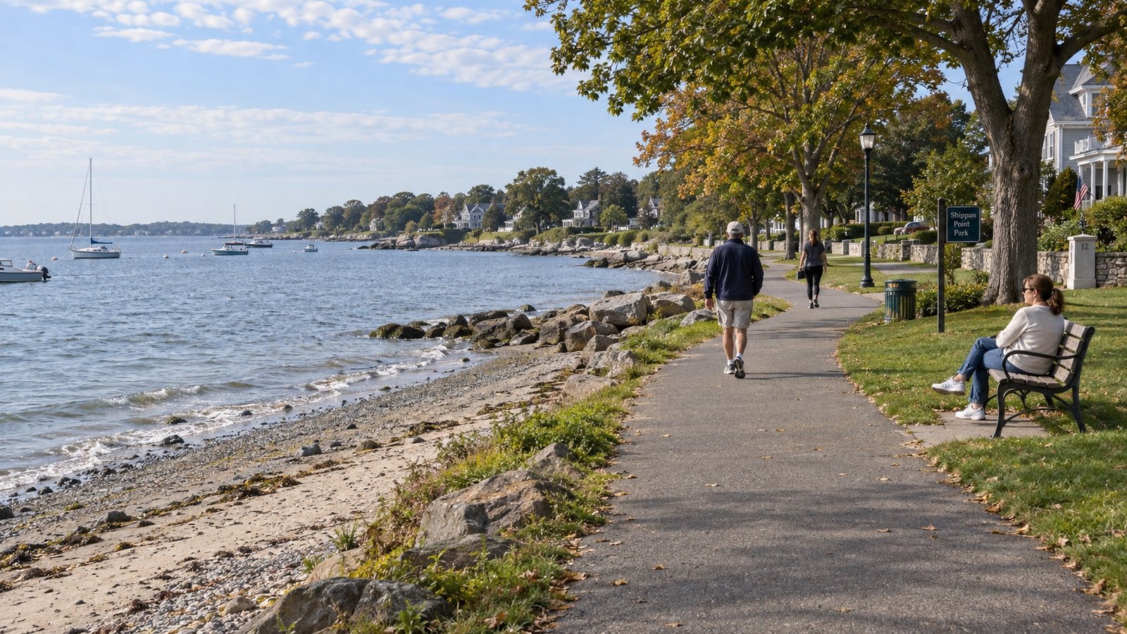 A maintained shoreline walking path beside small waves, pebbles and seaweed, with walkers and scattered boats.