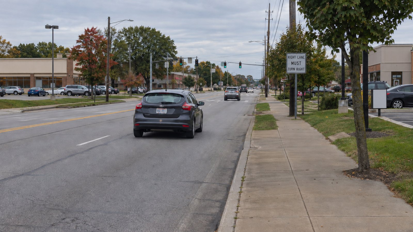 A whole hatchback travels toward traffic lights along a suburban road with a continuous sidewalk.
