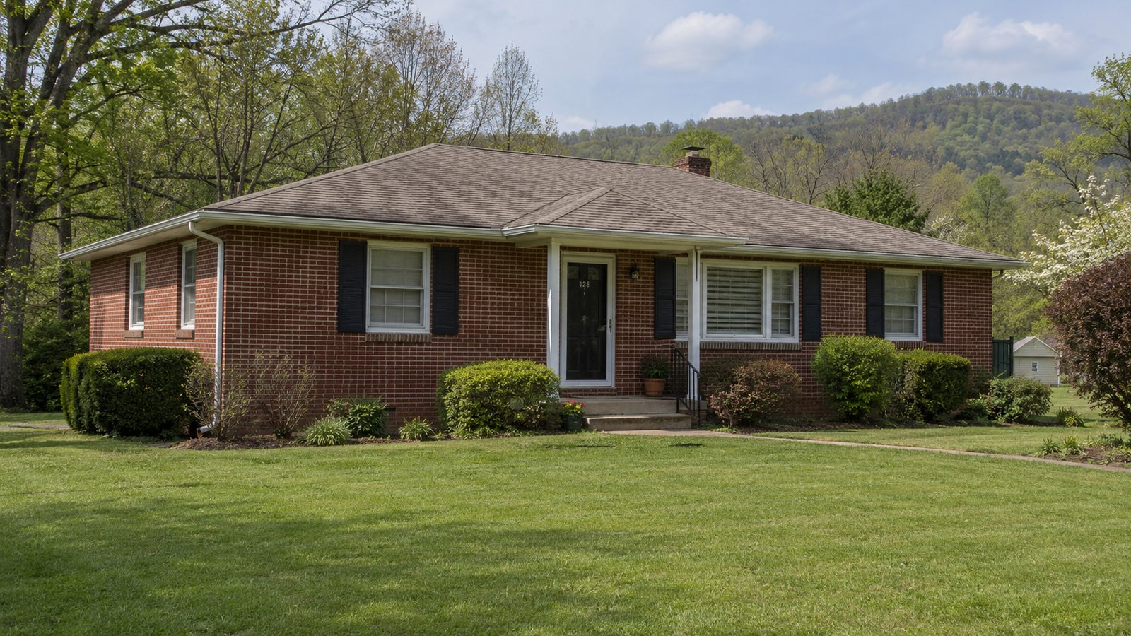 A home and its approach Single-story red-brick house with a low hip roof, front entrance, lawn and wooded hills behind