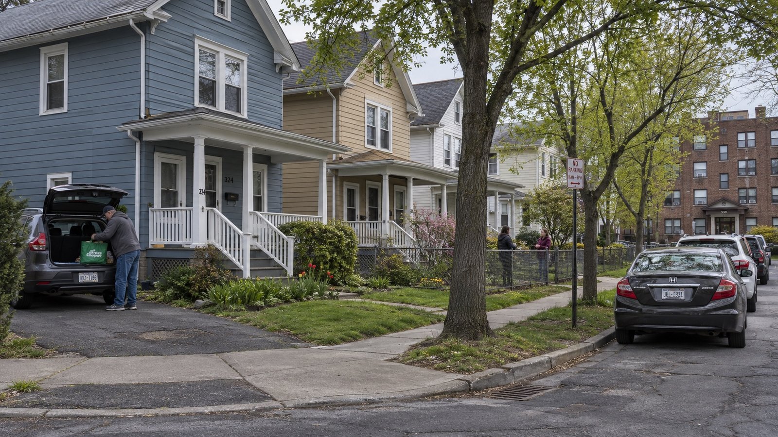 A residential street of older frame doubles with porches and parked cars.