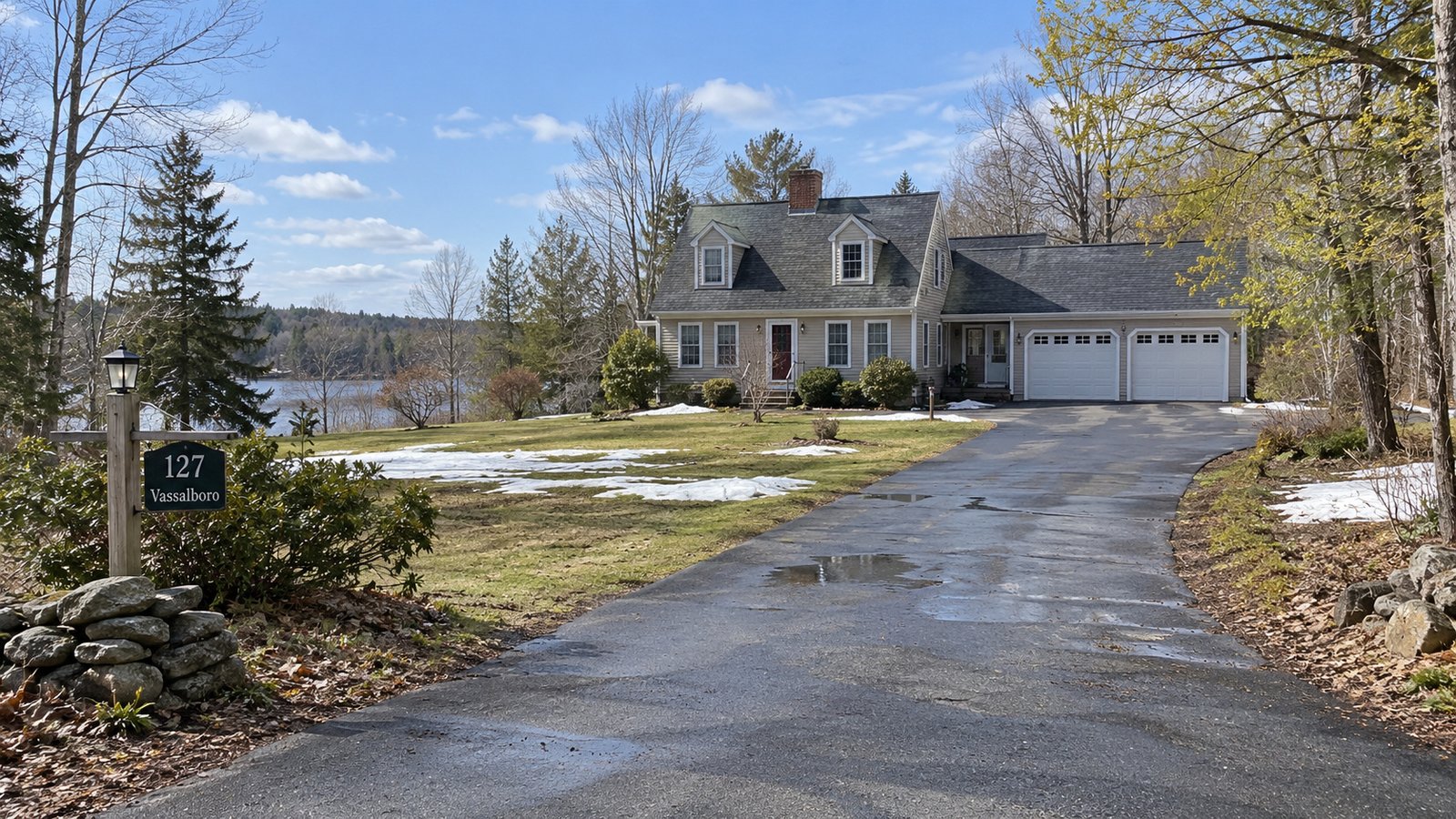 A complete country house and attached garage beside a connected damp driveway, with thawing snow patches in the yard under daylight.