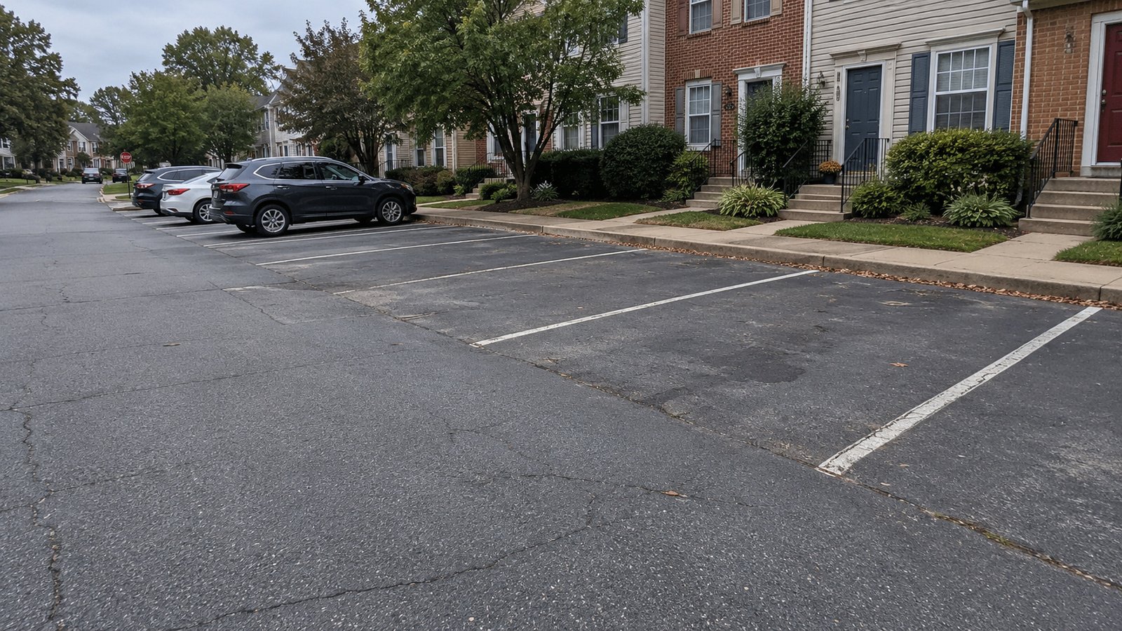 Review the open bays and the pedestrian route Two open asphalt car bays face townhome entrance paths beside a parking aisle that connects to a residential street.