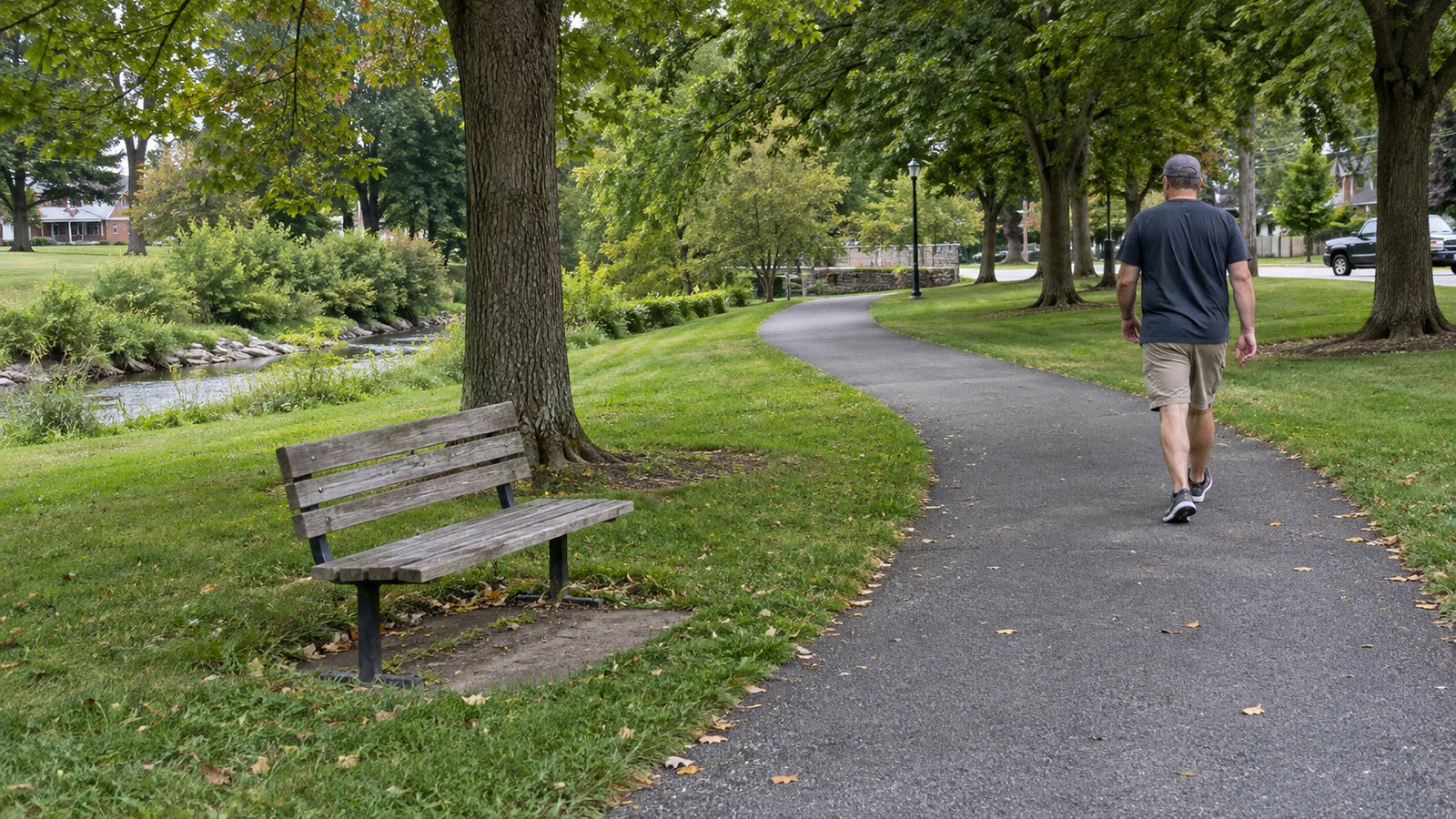 A man walking away on a curving park path beside a wooden bench and mature trees.
