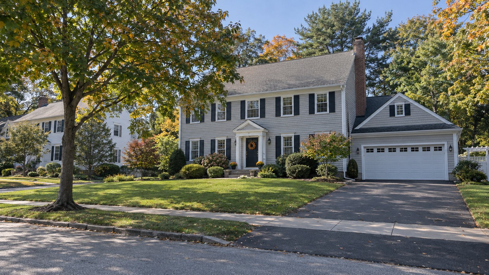 Reviewing a residential approach Colonial-style house with a garage, driveway and mature trees.