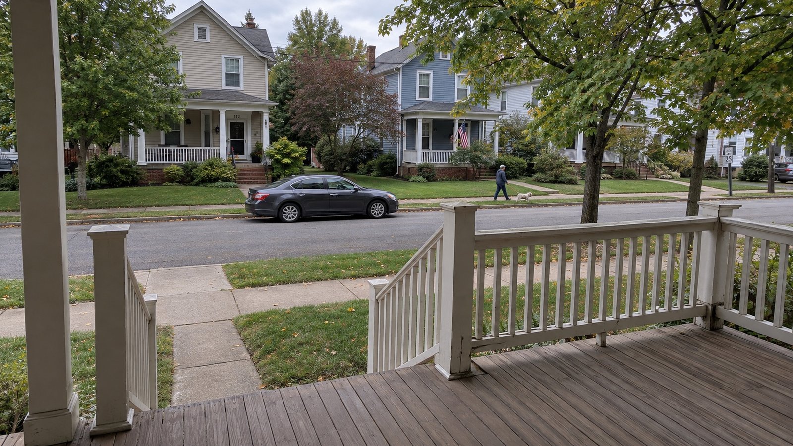 View from a raised wooden porch across a sidewalk toward older houses and a resident walking a dog