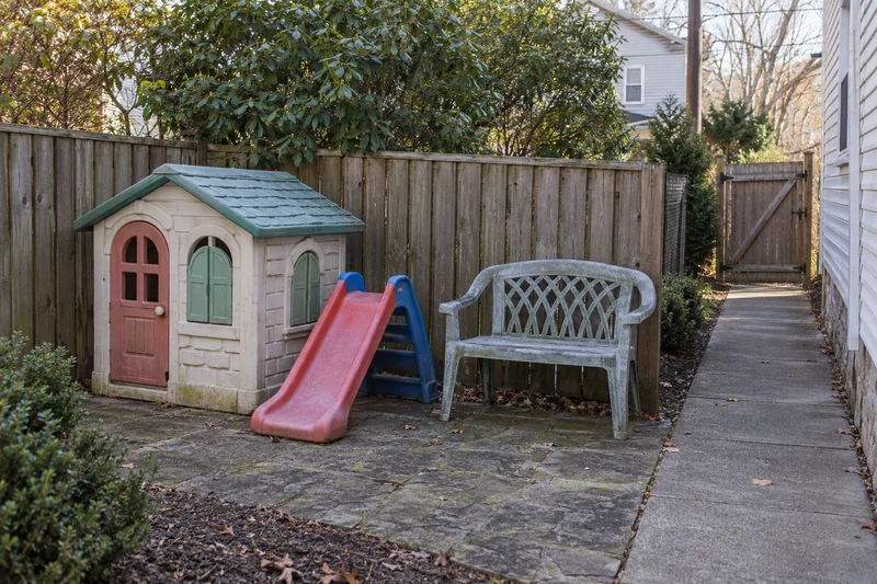 Portable playhouse, low slide and faded resin bench beside a clear side path Outgrown portable play items and a garden bench grouped beside a clear private gate