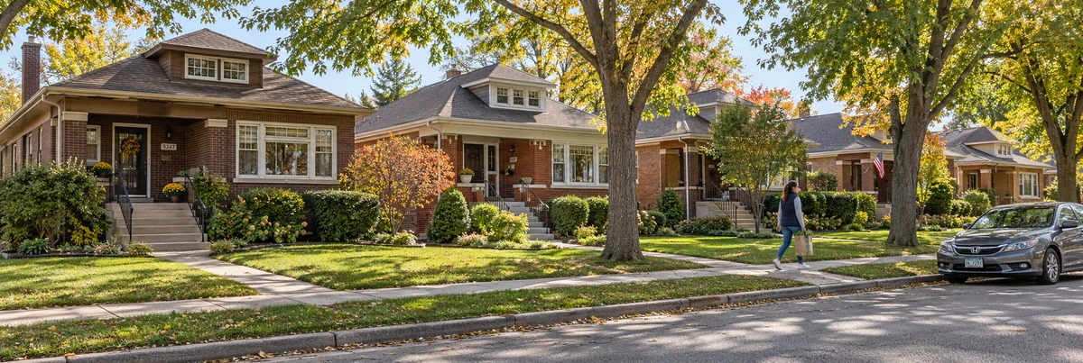 An adult carrying a grocery bag walks along a sidewalk beside brick bungalows and autumn trees.