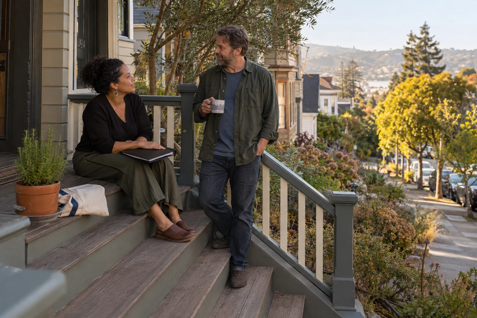 An Oakland evening around work and learning A woman rests a closed dark folder on her lap while a man holds a ceramic mug beside wooden front steps overlooking a sloping residential street.