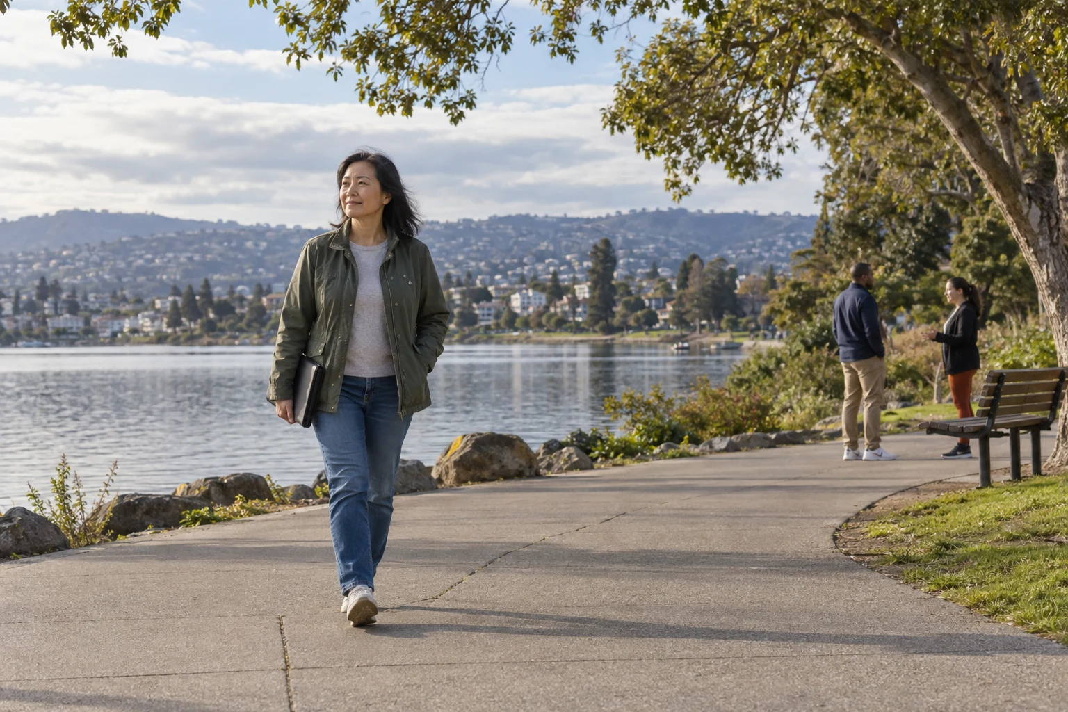 An Oakland lakeside outing Woman carrying a plain portfolio walks along a broad urban lakeside path while two other adults talk near a bench.